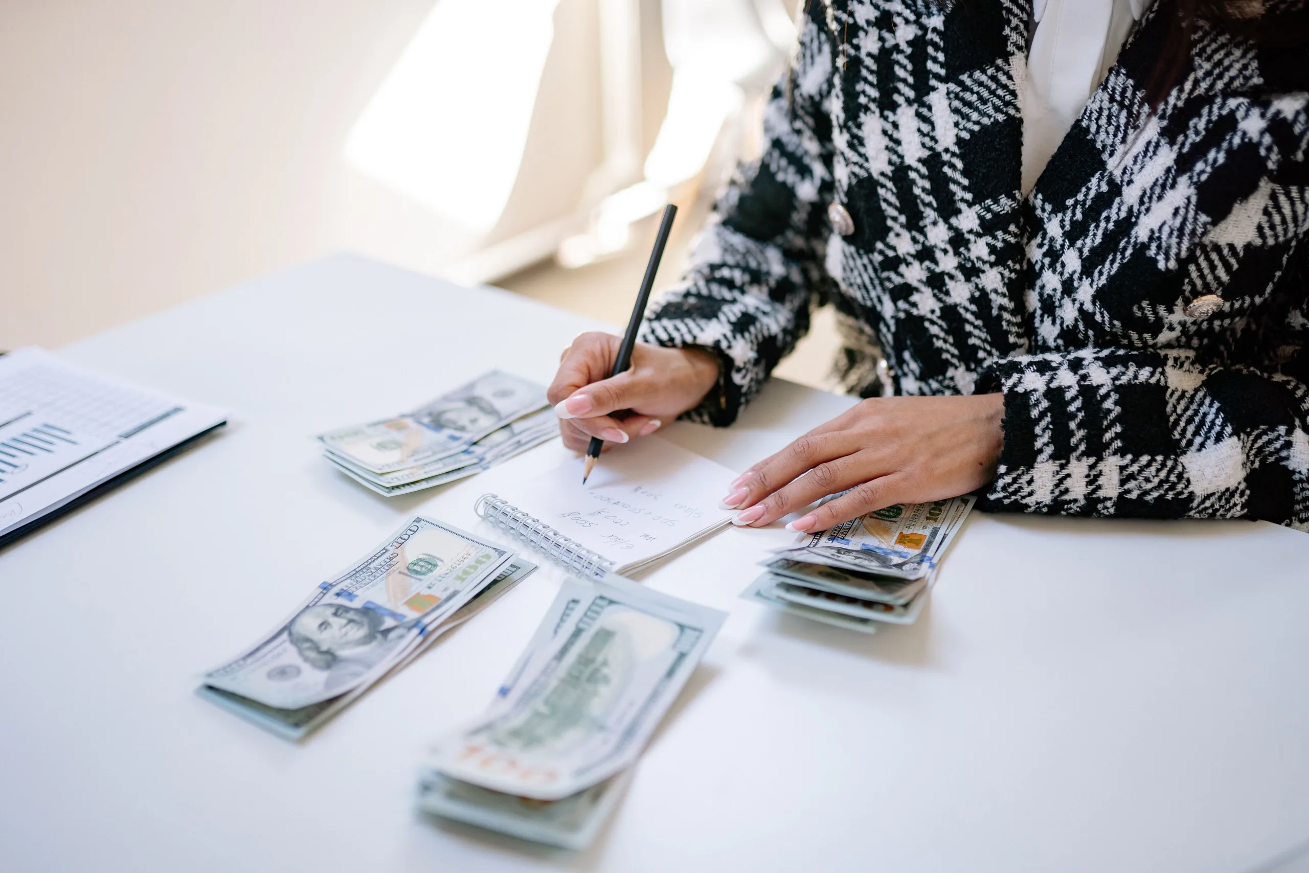 Bureau d’une PME vu de dessus : ordinateur ouvert sur un tableur, dossiers de factures, calculatrice, et une dirigeante qui note des montants dans un carnet